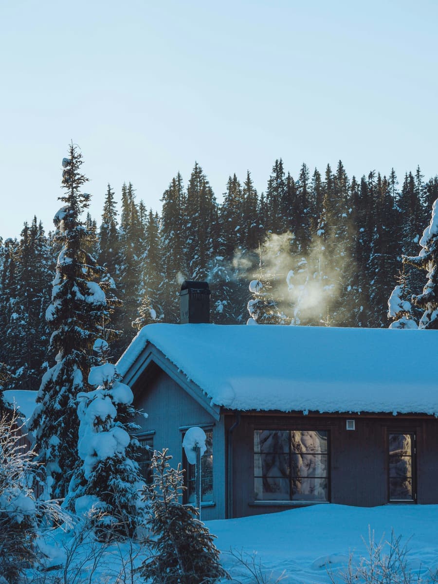 A snow-covered house in the woods with smoke rising from its chimney.